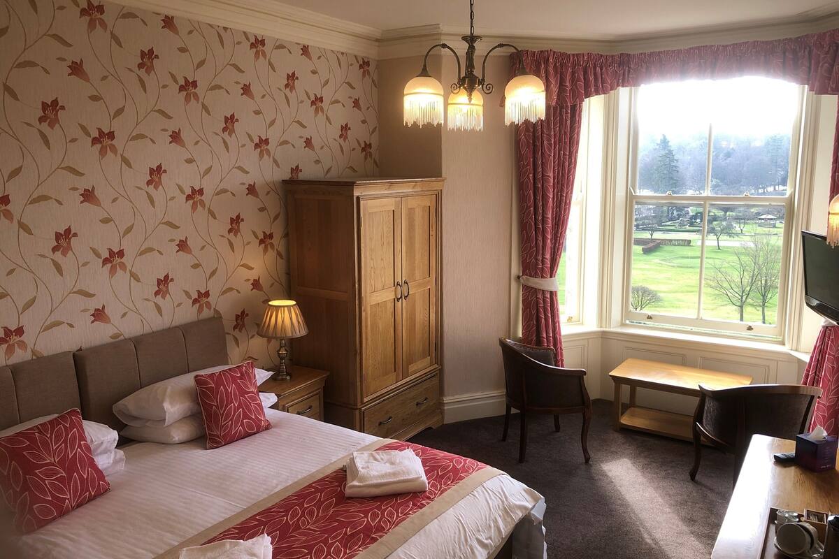 Une chambre d’hôtel avec un grand lit, une armoire en bois, un bureau avec une chaise et une fenêtre donnant sur un parc.
