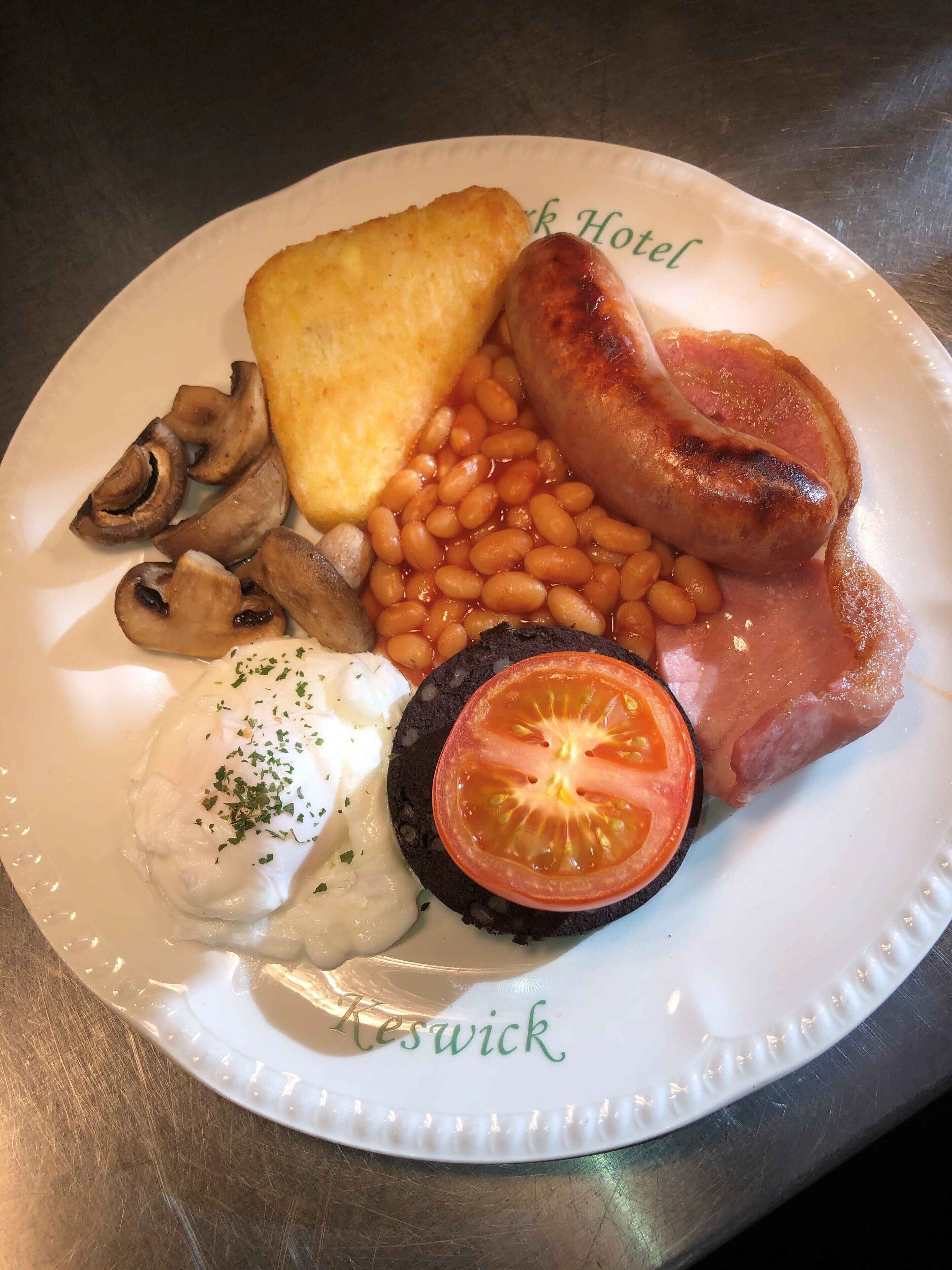A plate of breakfast food including sausages, beans, mushrooms, toast, ham, and a slice of tomato.