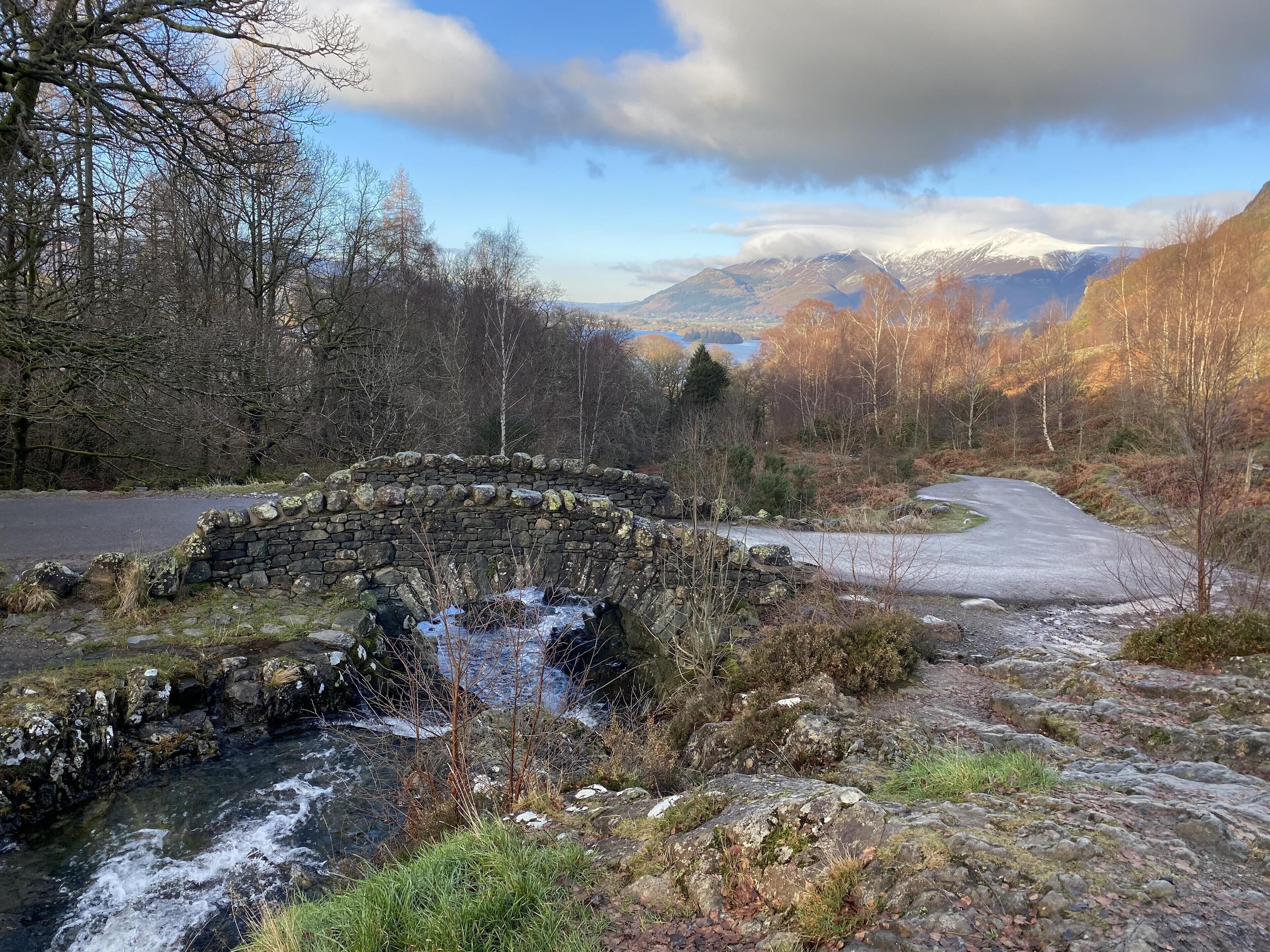 A stone bridge over a stream in a forested area with a road curving around it and mountains in the background.