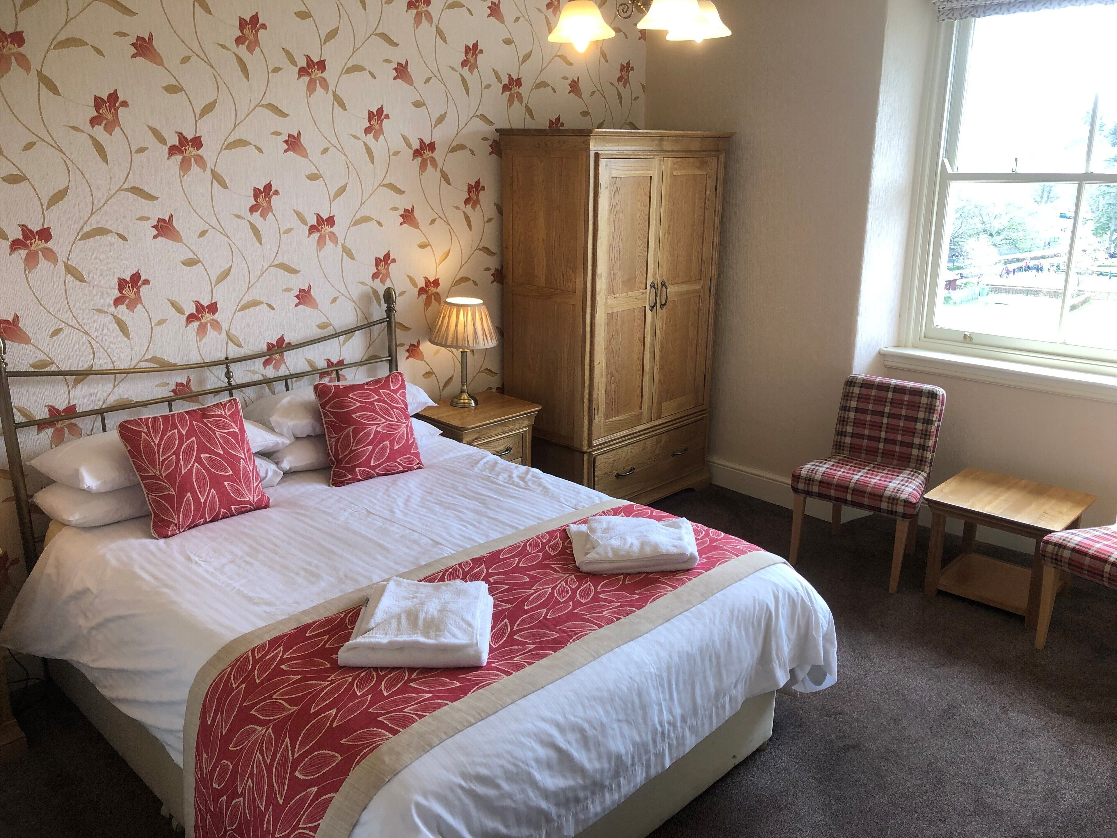 A neatly made bed with red and white bedding, a wooden wardrobe, a plaid chair, and a small table.