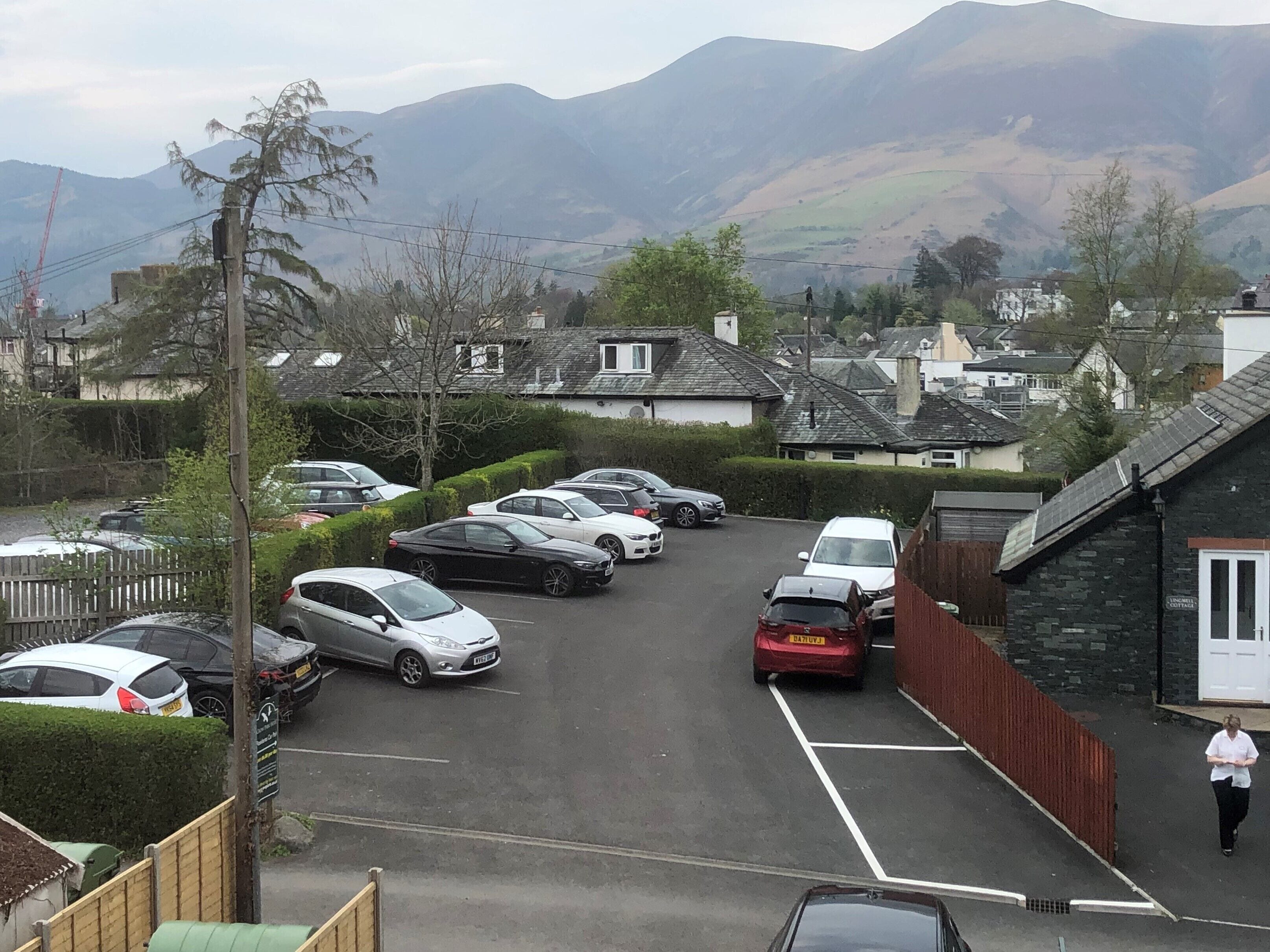 A parking lot with several cars, a person walking, and houses in the background with mountains.