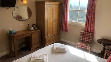 A bedroom with a bed, a wooden wardrobe, a chair, a small table, a mirror, and a window with curtains.