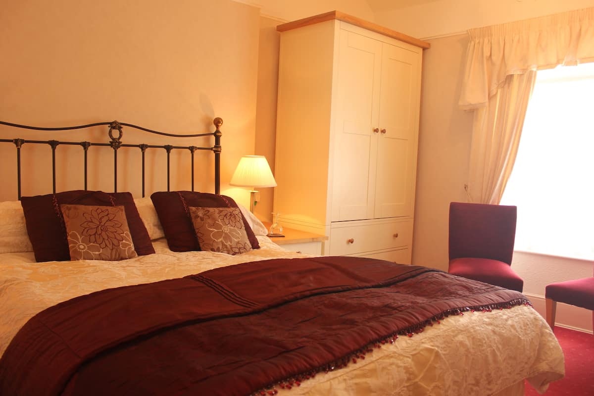 A neatly made bed with a dark brown bedspread, a white and brown patterned pillow, and a wooden headboard. There is a white nightstand with a lamp and a red chair next to a window with curtains.