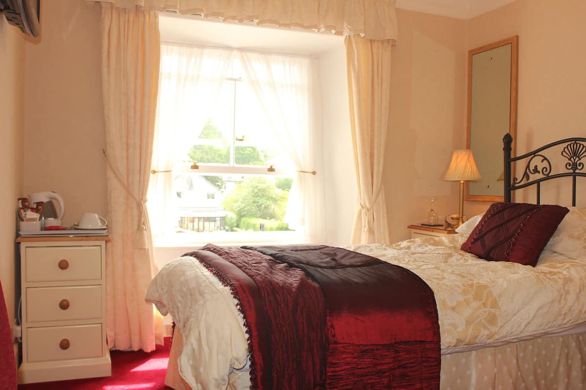 A bedroom with a bed, a nightstand, a dresser, a window with curtains, and a mirror on the wall.