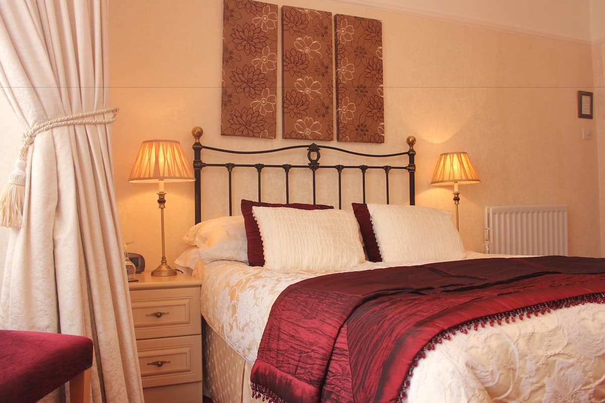 A neatly made bed with a red and white bedspread, two bedside lamps, a nightstand with a lamp, and a wall with three floral paintings.