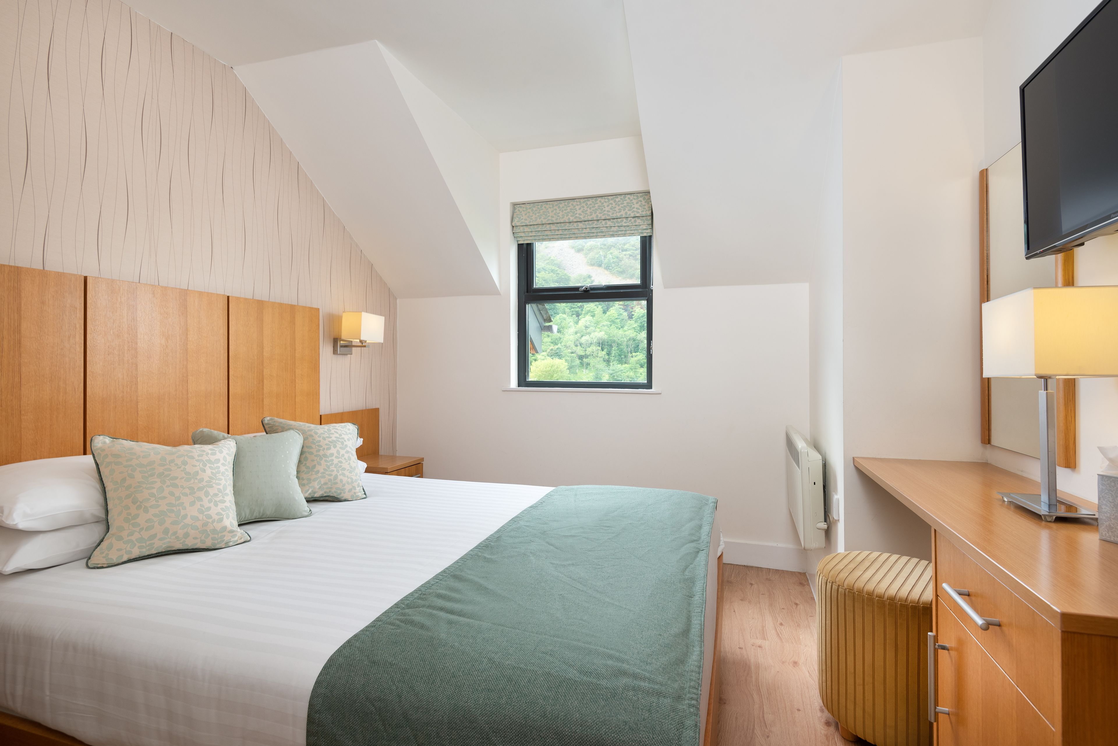 A bedroom with a bed, bedside lamps, a window, a radiator, and a TV.