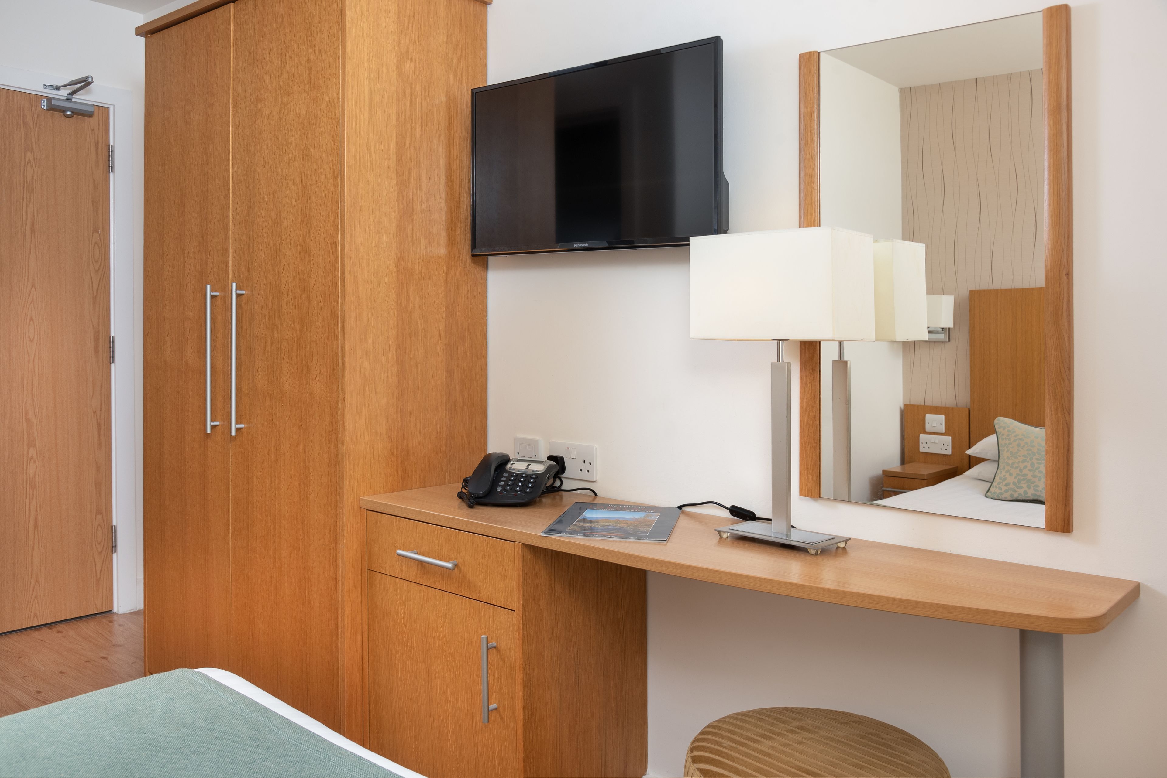 A hotel room with a wooden wardrobe, a desk with a lamp, a flat-screen TV, and a bed with a green bedspread.