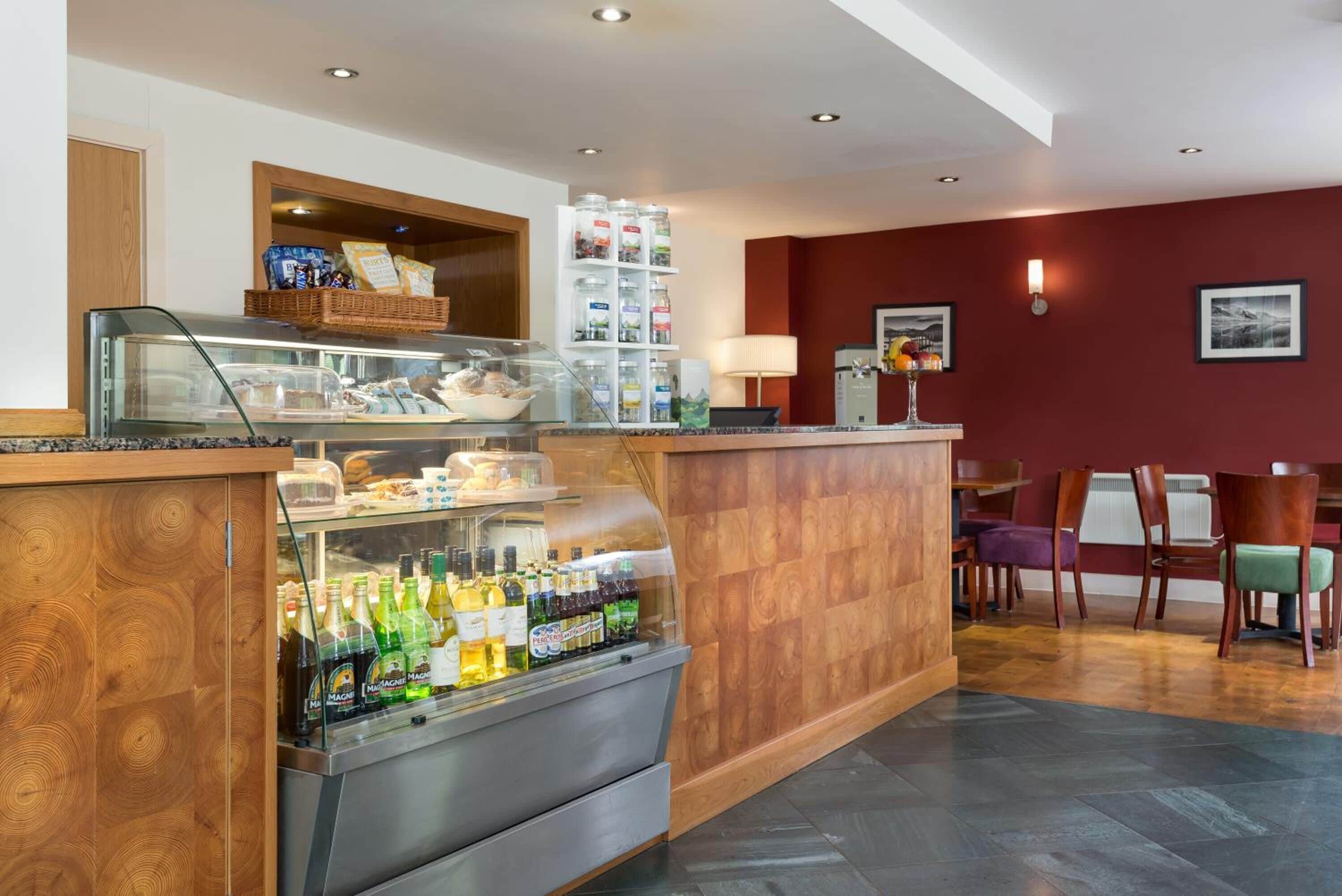 A hotel lobby with a wooden reception desk, a glass display case with snacks, a beverage cooler, and a seating area with chairs and a table.