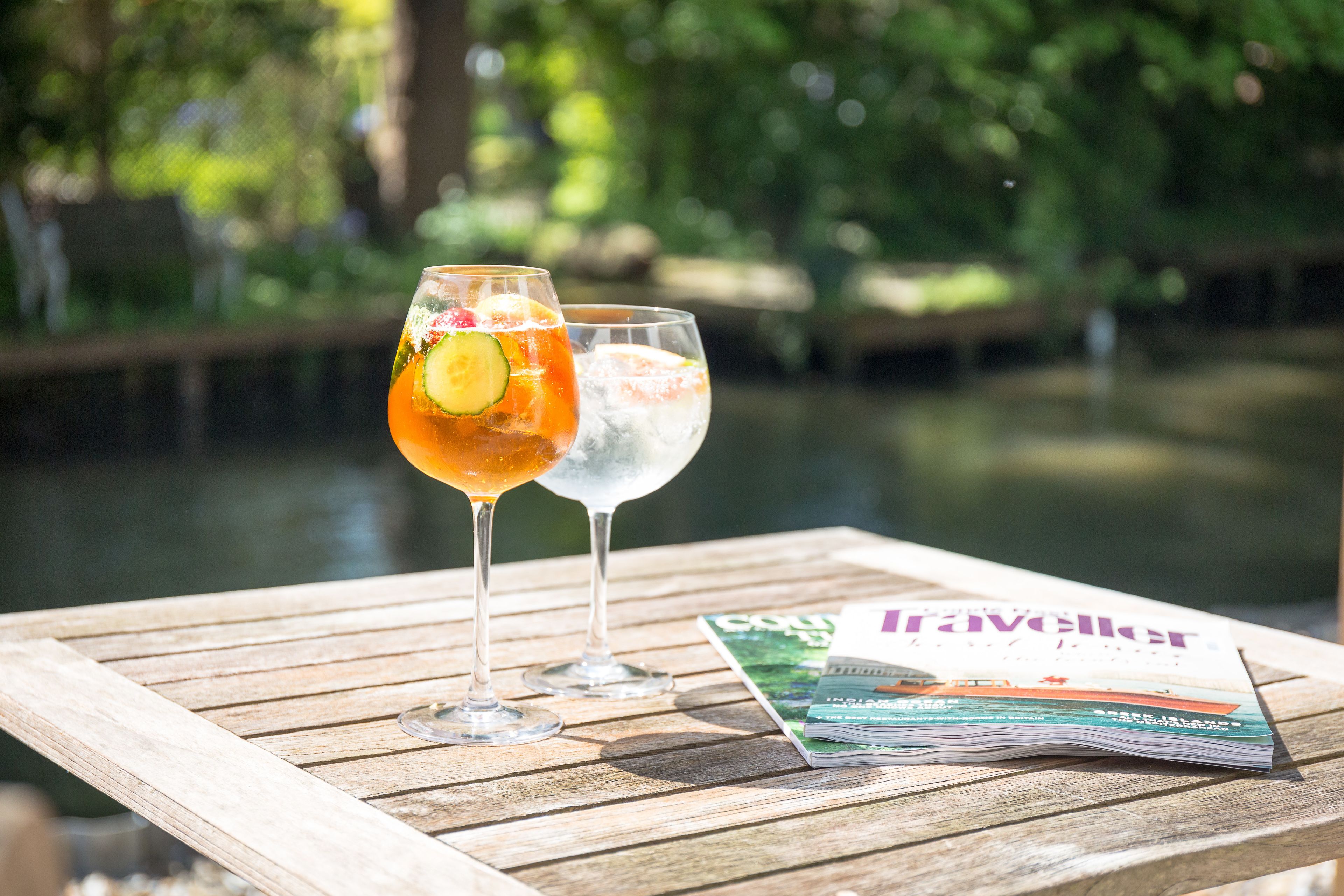 Two glasses of beverages on a wooden table with a magazine, set against a blurred natural background.
