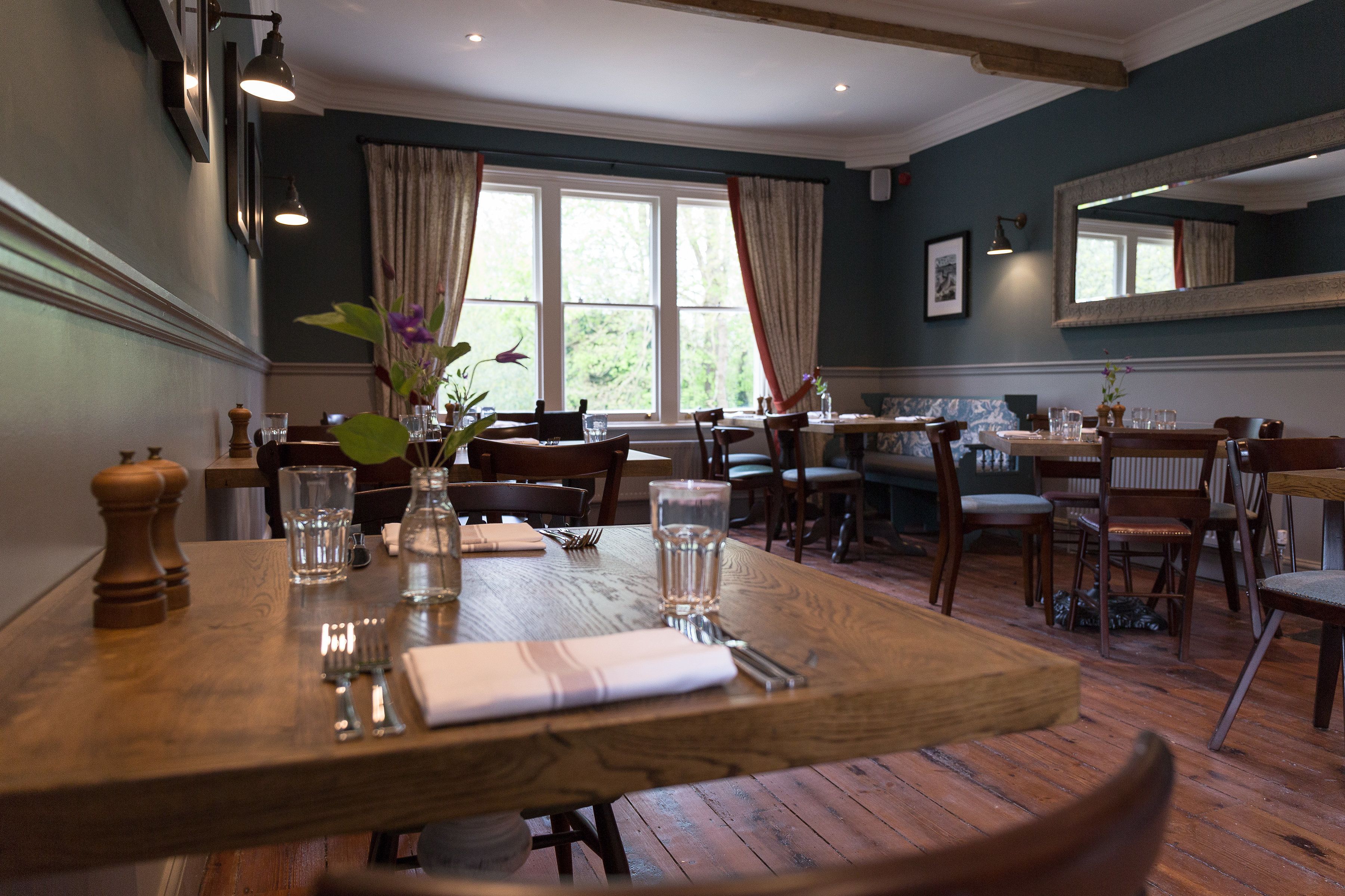 A dining area with wooden tables and chairs, large windows with curtains, and a mirror on the wall.