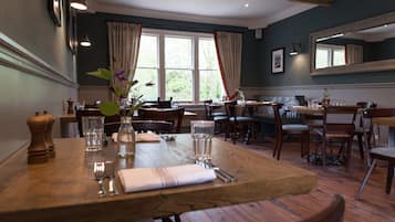 A dining area with wooden tables and chairs, large windows with curtains, and a mirror on the wall.