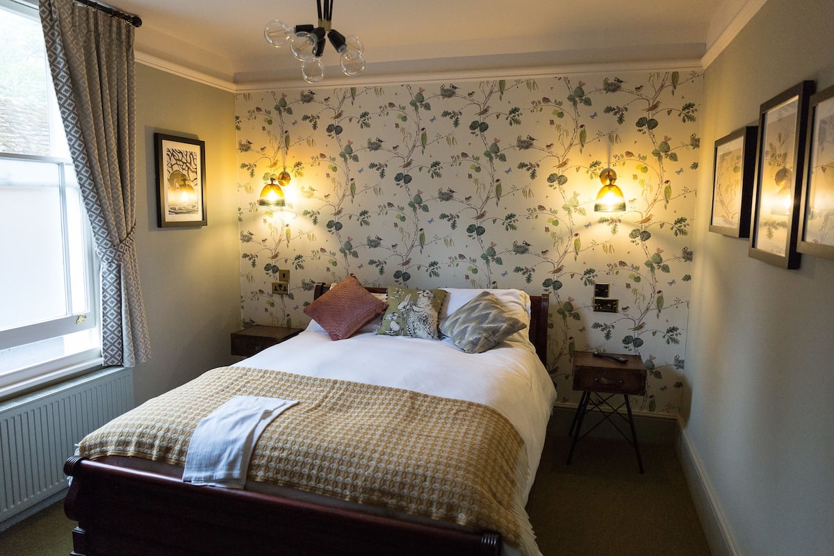 A neatly made bed with a patterned bedspread, two wall-mounted reading lamps, and a window with curtains.