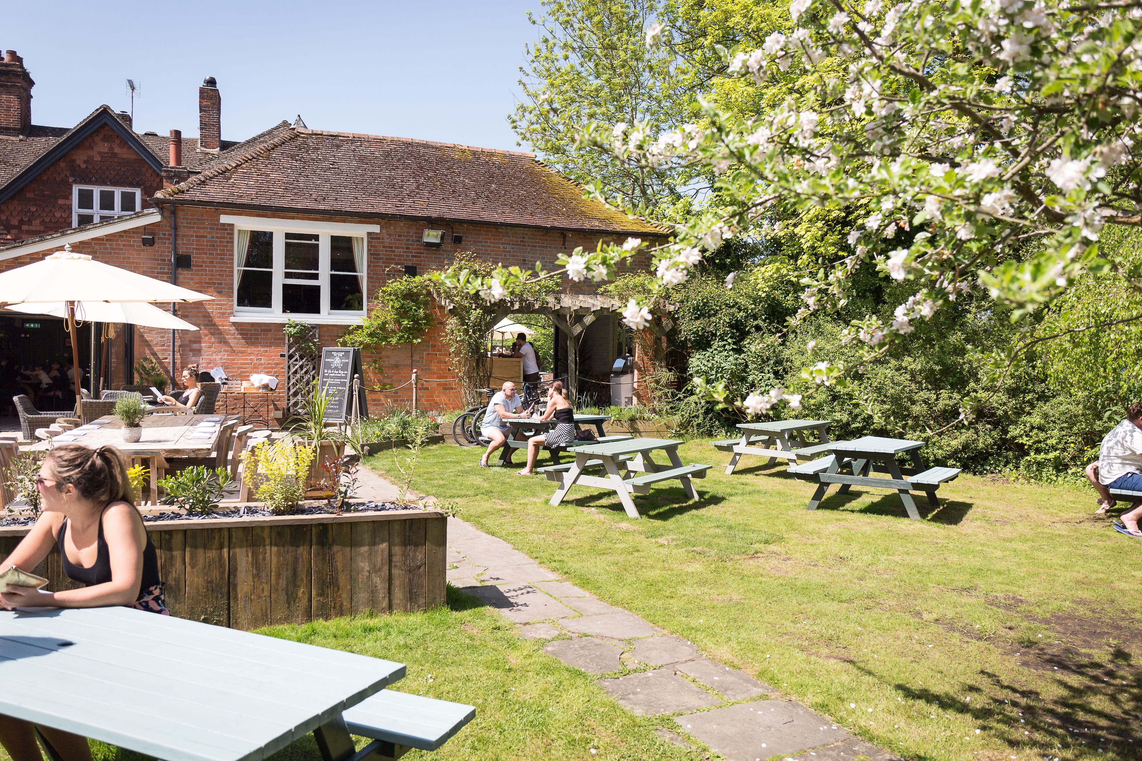 An outdoor seating area with tables and chairs, a brick building, and a garden with flowering trees.