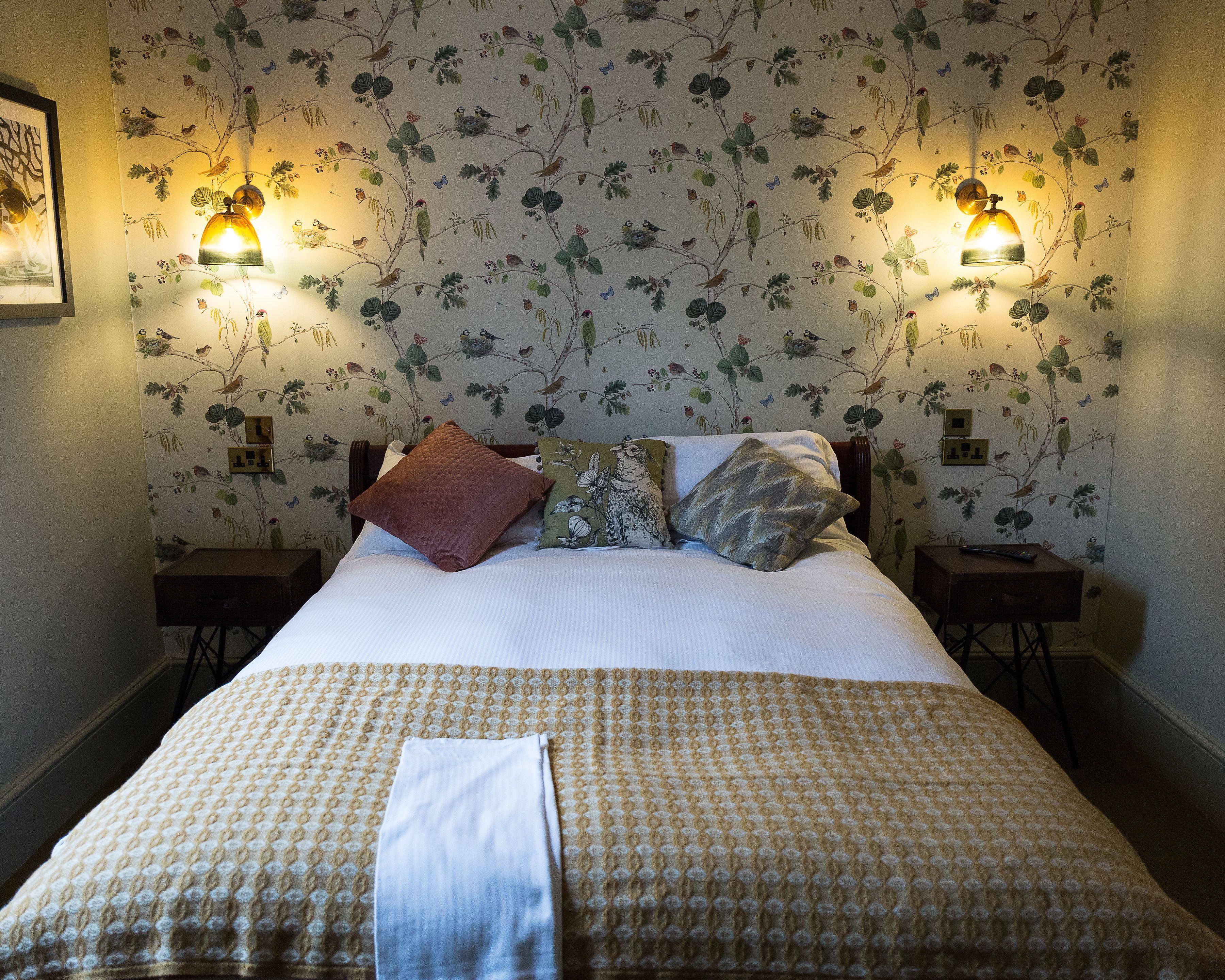 Un lit bien fait, recouvert d’une couvre-lit à motifs, deux tables de chevet avec des lampes et un papier peint à fleurs.
