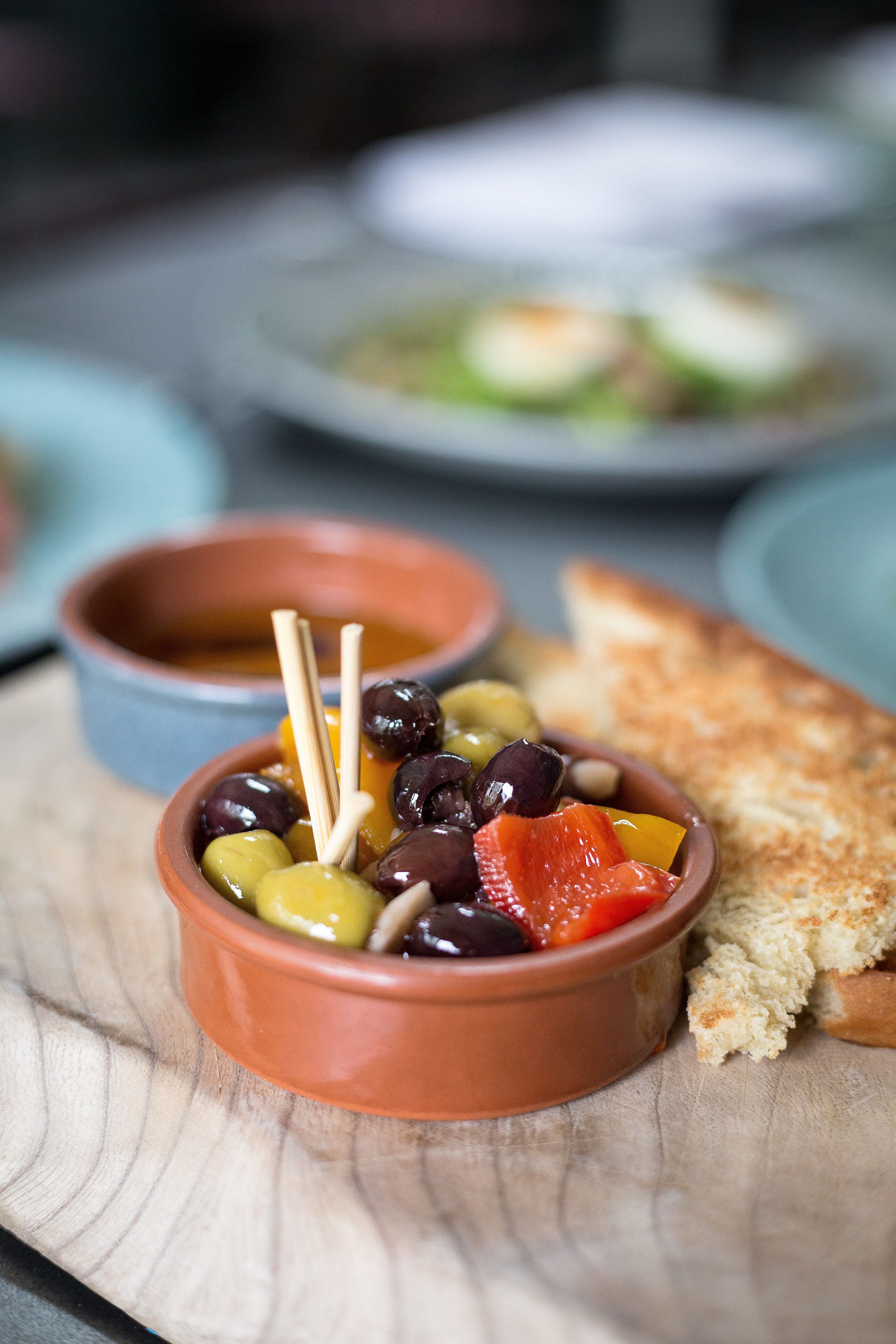 A bowl of olives, beans, and tomatoes on a wooden board with a piece of bread.