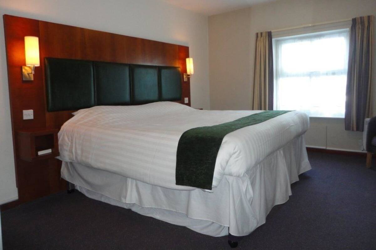 A hotel room with a large bed, wooden headboard, two wall-mounted lamps, a window with curtains, and a carpeted floor.