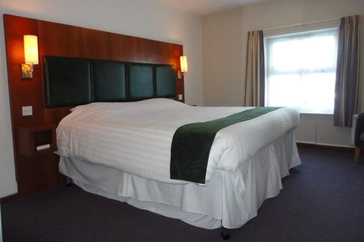 A hotel room with a large bed, wooden headboard, two wall-mounted lamps, a window with curtains, and a carpeted floor.
