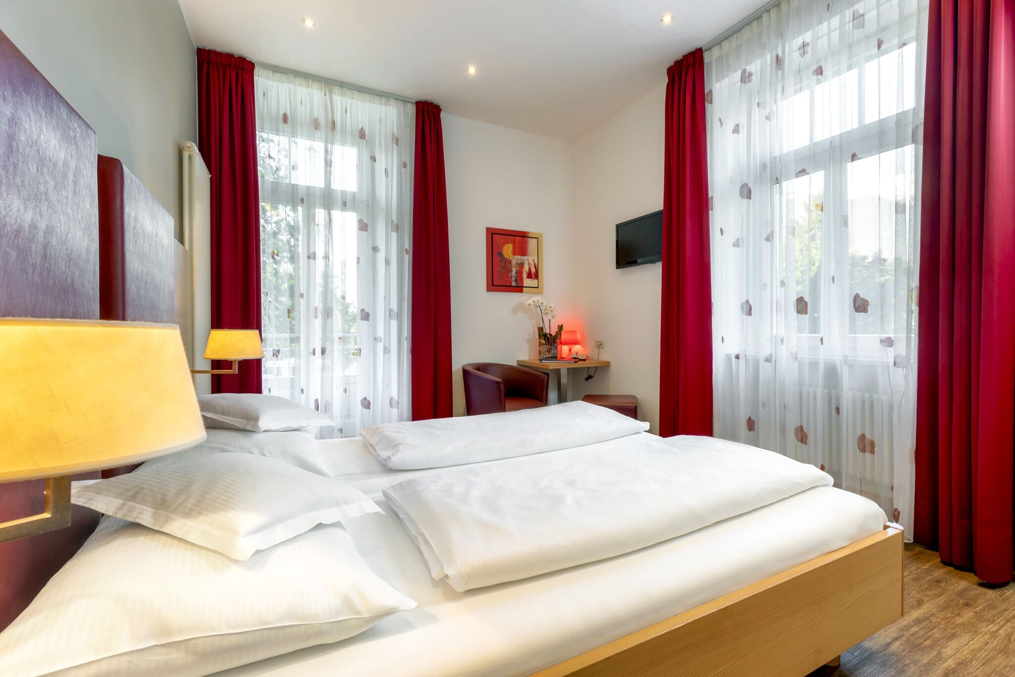 A hotel room with a bed, a desk, a chair, a TV, and a window with red curtains.