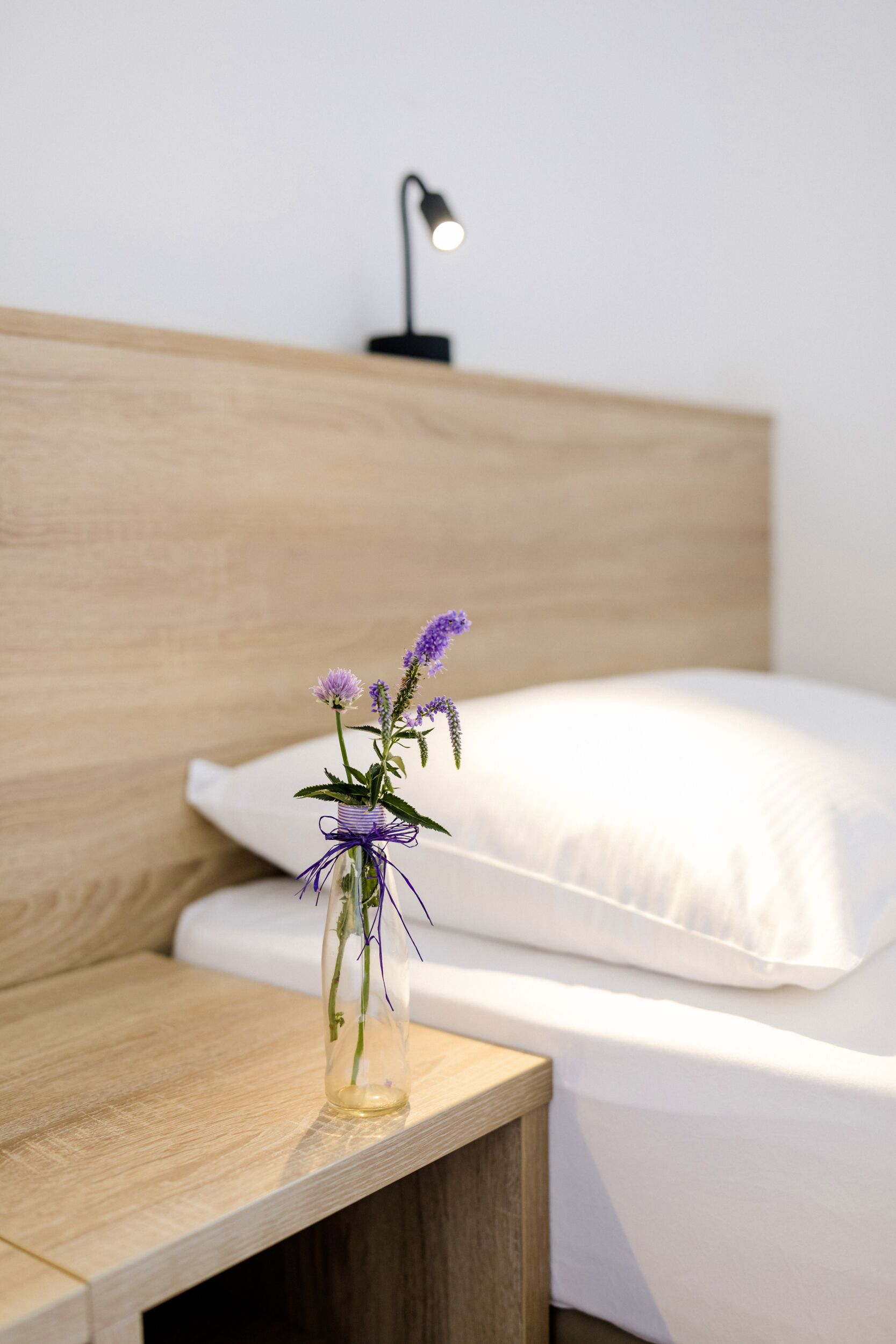 Een netjes opgemaakt bed met witte lakens en een houten hoofdbord. Een klein tafeltje naast het bed heeft een glazen vaas met paarse bloemen.
