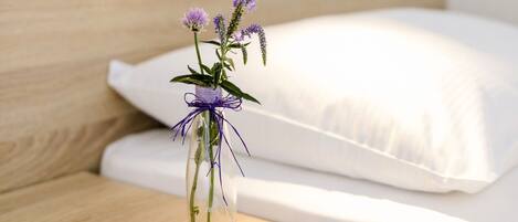Ein ordentlich bezogenes Bett mit weißen Laken und einem hölzernen Kopfteil. Ein kleiner Tisch daneben trägt eine transparente Vase mit lila Blumen.
