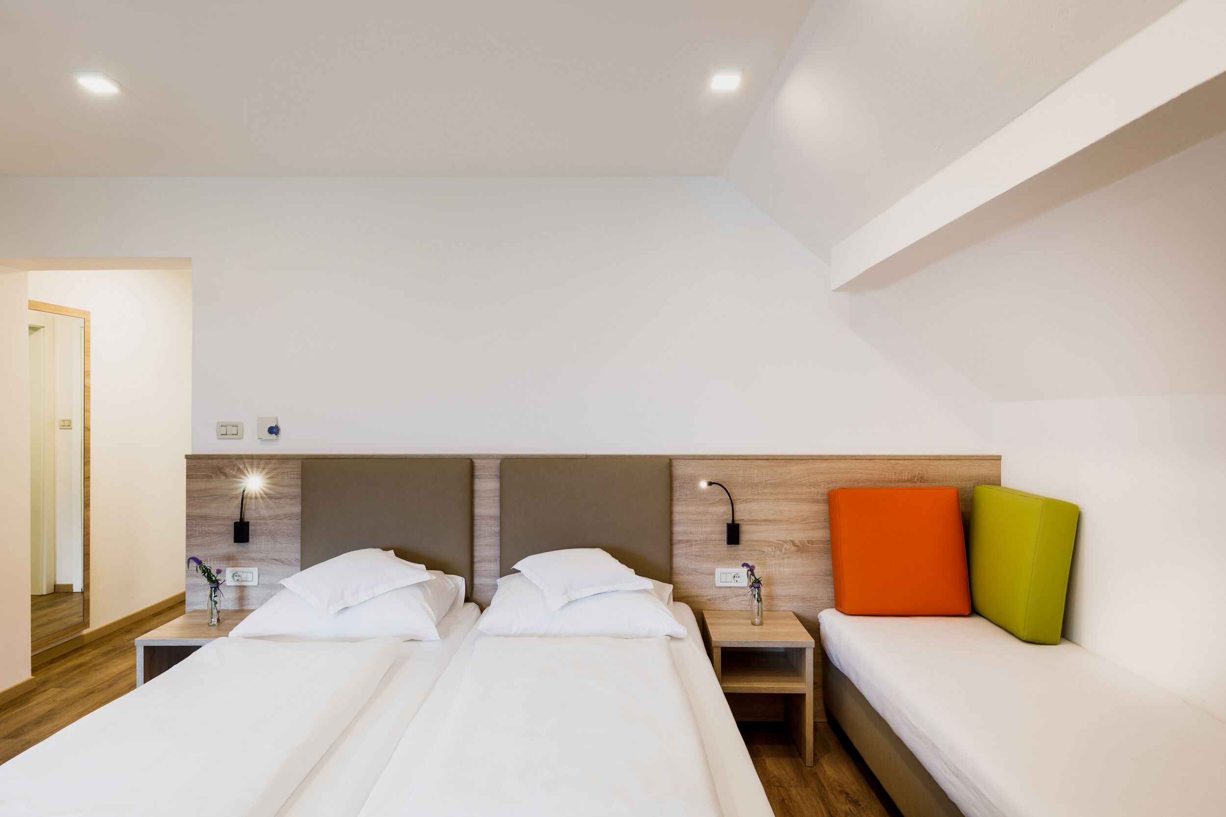 Quarto de hotel com duas camas, uma mesa de cabeceira com abajur e luminária na parede.