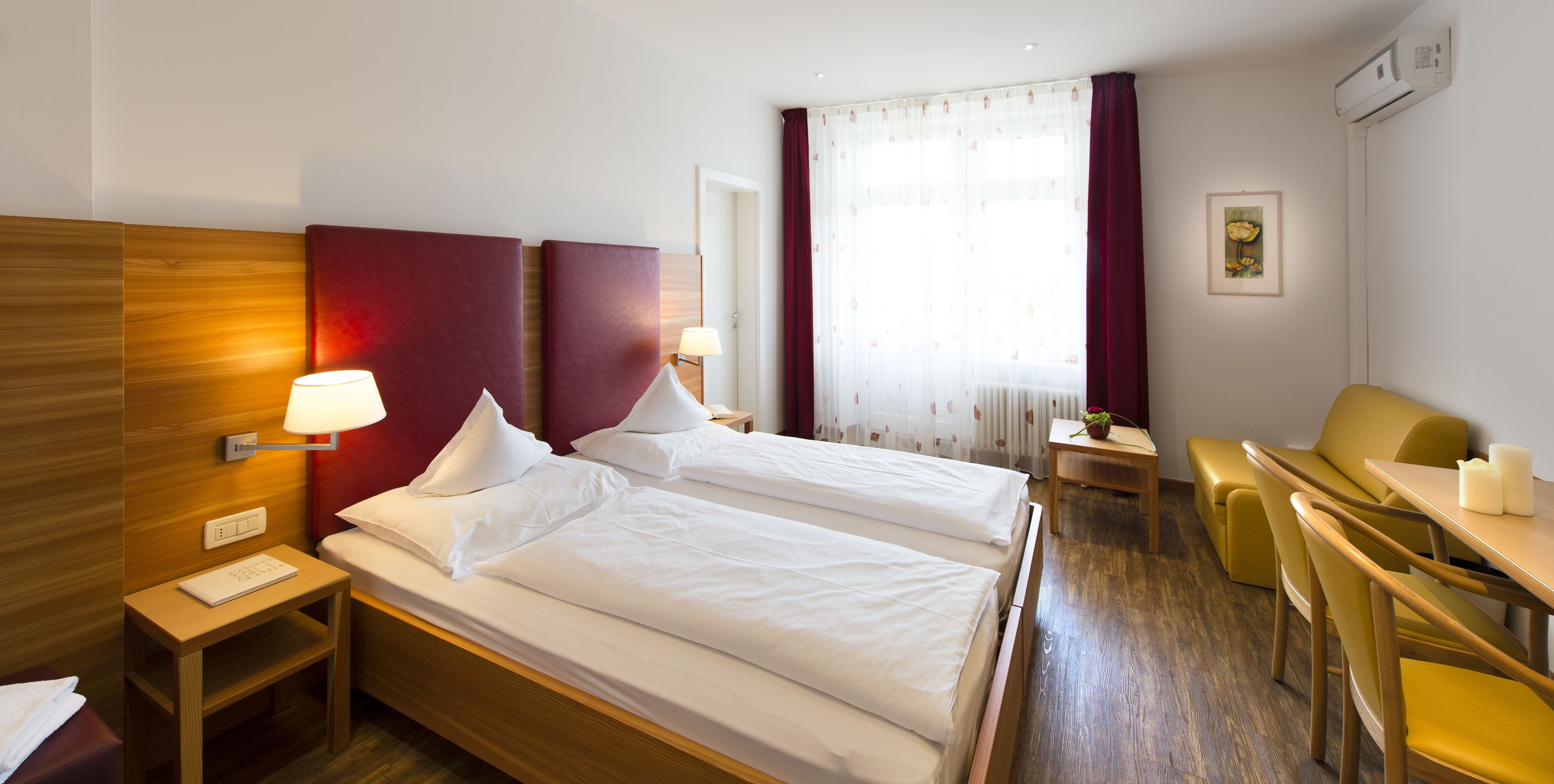 A hotel room with two single beds, a desk with a chair, a small table with a lamp, and a window with curtains.