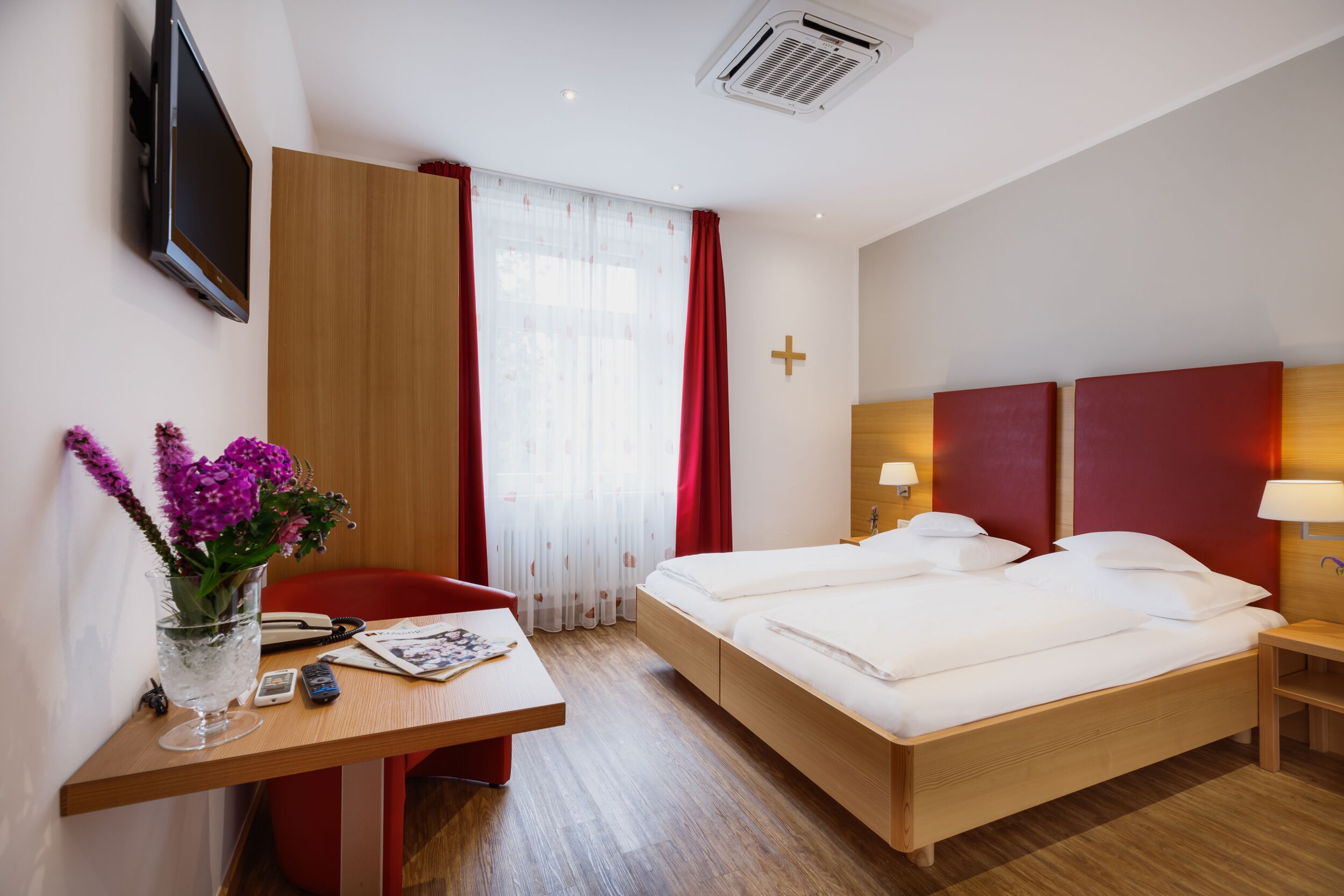 Ein Hotelzimmer mit einem Bett, einem Schreibtisch mit Blumen in einer Vase, einem Fernseher und einem Kreuz an der Wand.
