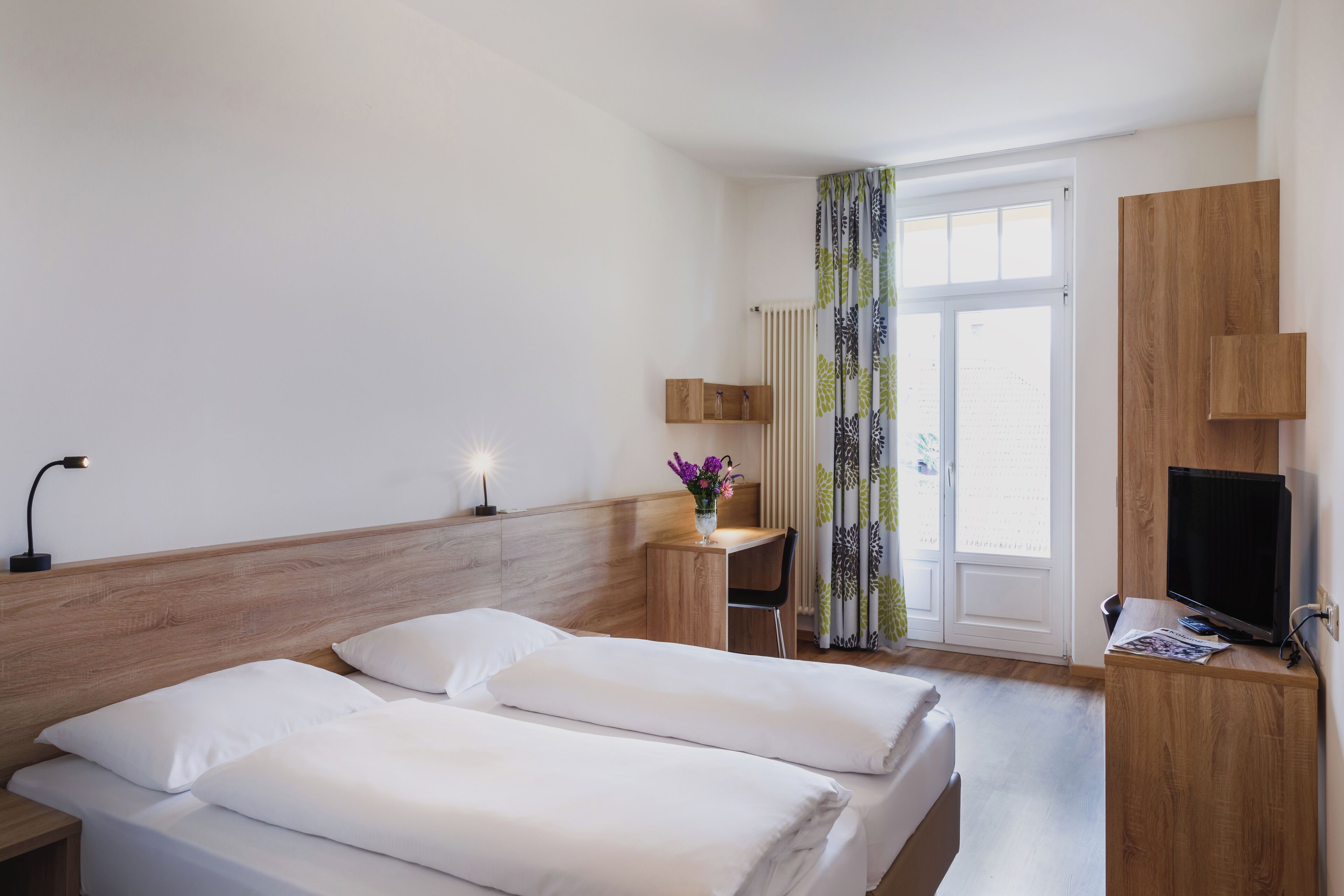 Een hotelkamer met een bed, nachtkastje, lamp, televisie en een raam met gordijnen.