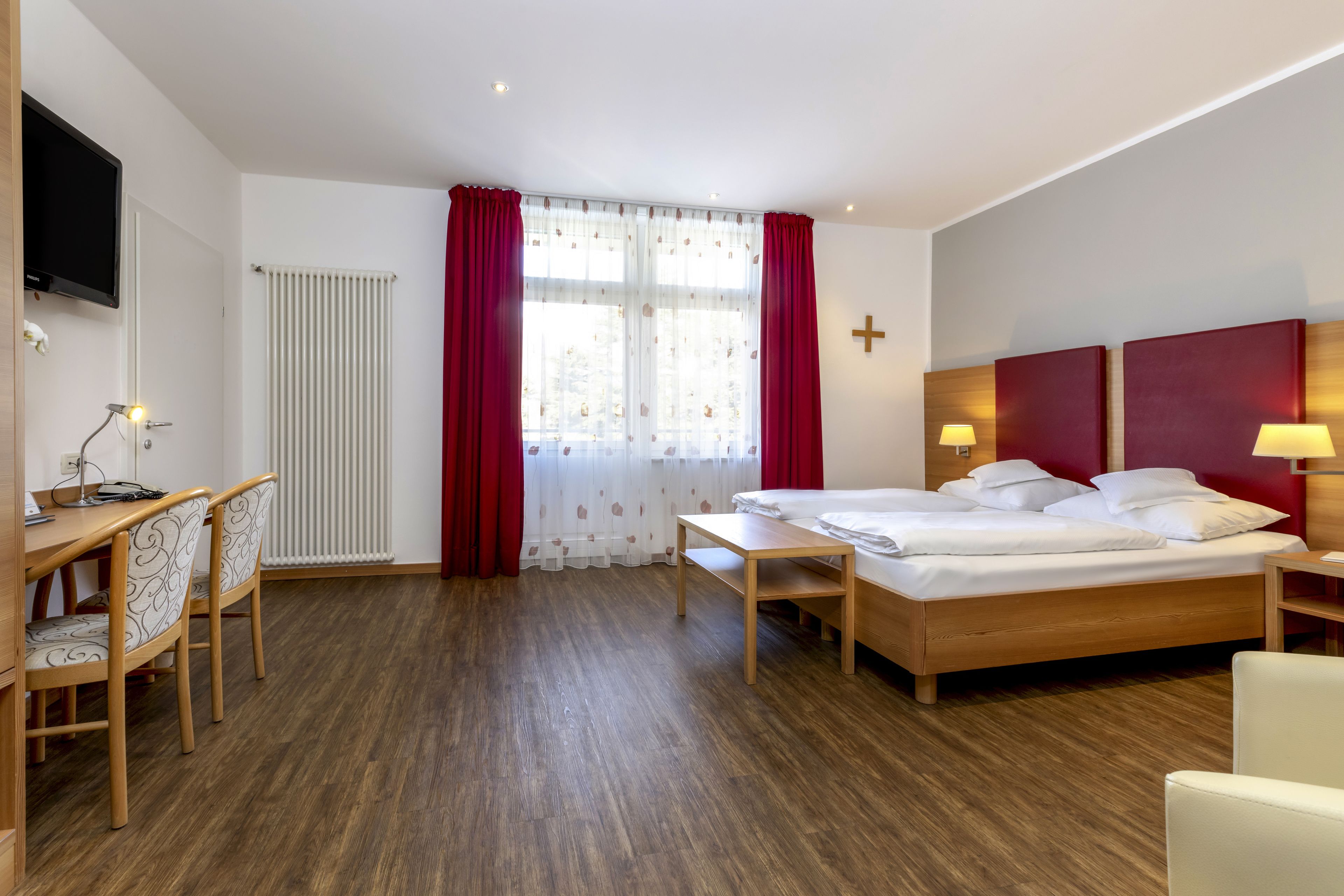 A hotel room with a wooden bed, a desk with a chair, a TV mounted on the wall, and a window with red curtains.