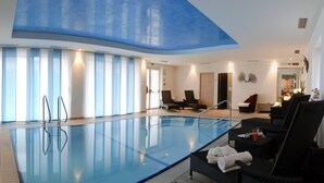 Piscina interna moderna com paredes de vidro transparente, cadeiras de descanso e teto com design azul.