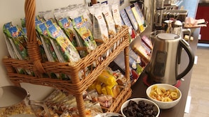 Uma mesa de buffet com diversos alimentos e acompanhamentos, incluindo uma cafeteira, tigelas de frutas e lanches embalados.