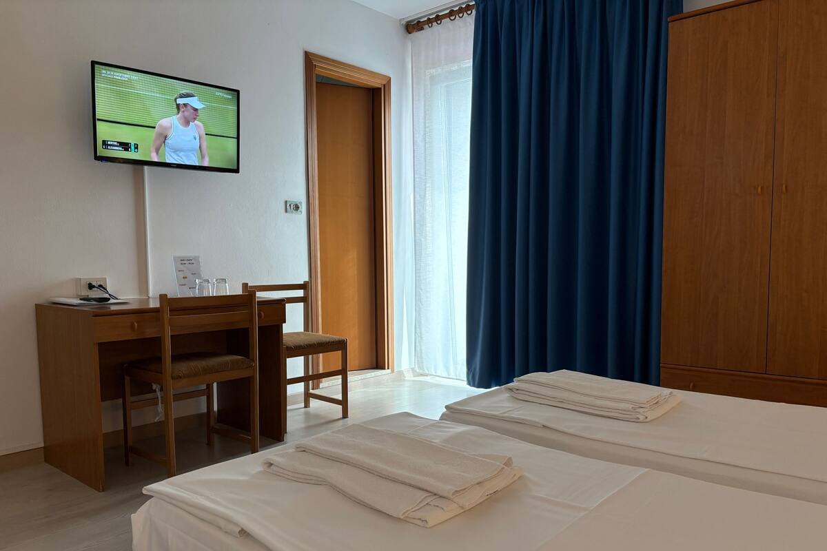 A hotel room with a bed, a desk with a chair, a television on the wall, and a wardrobe.
