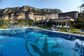 Una piscina con sillas de descanso, montañas al fondo y un edificio con balcones.