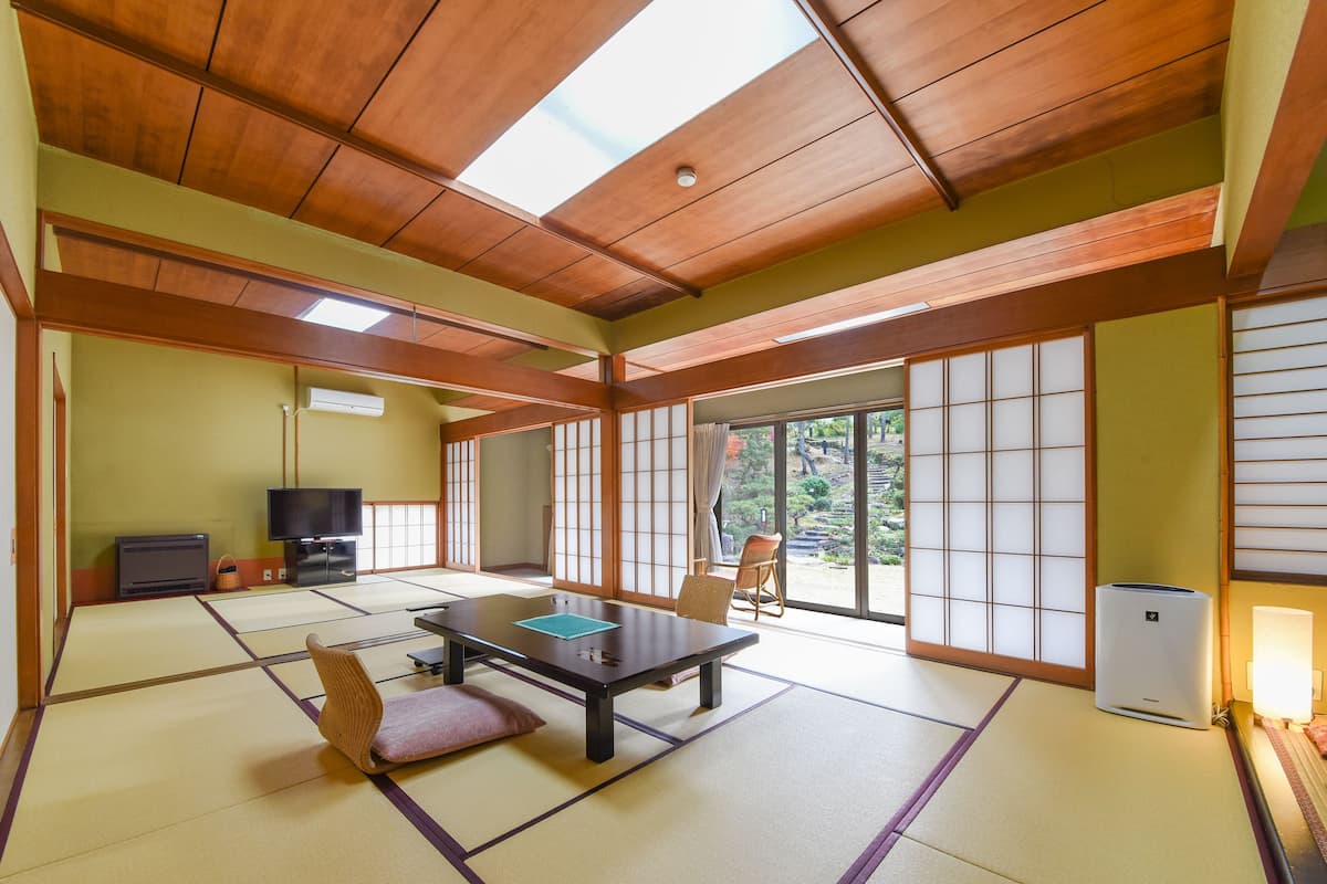 Ein traditioneller japanischer Raum mit Tatami-Bodenbelag, einem niedrigen Tisch, Stühlen, einem Fernseher und Schiebetüren, die zu einem Garten führen.