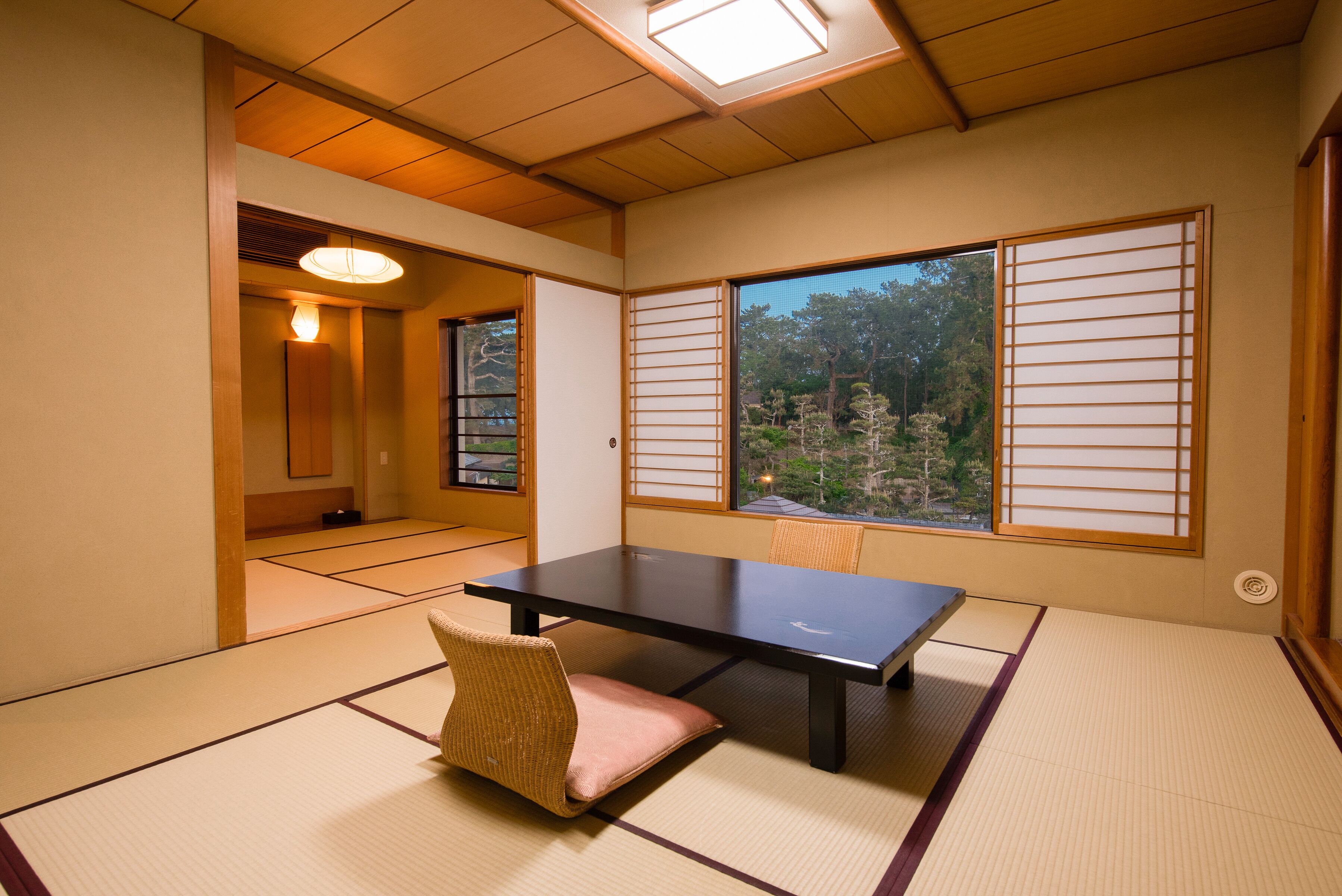 A tatami mat room with a low table and a cushioned chair, large windows with shoji screens, and a view of trees.