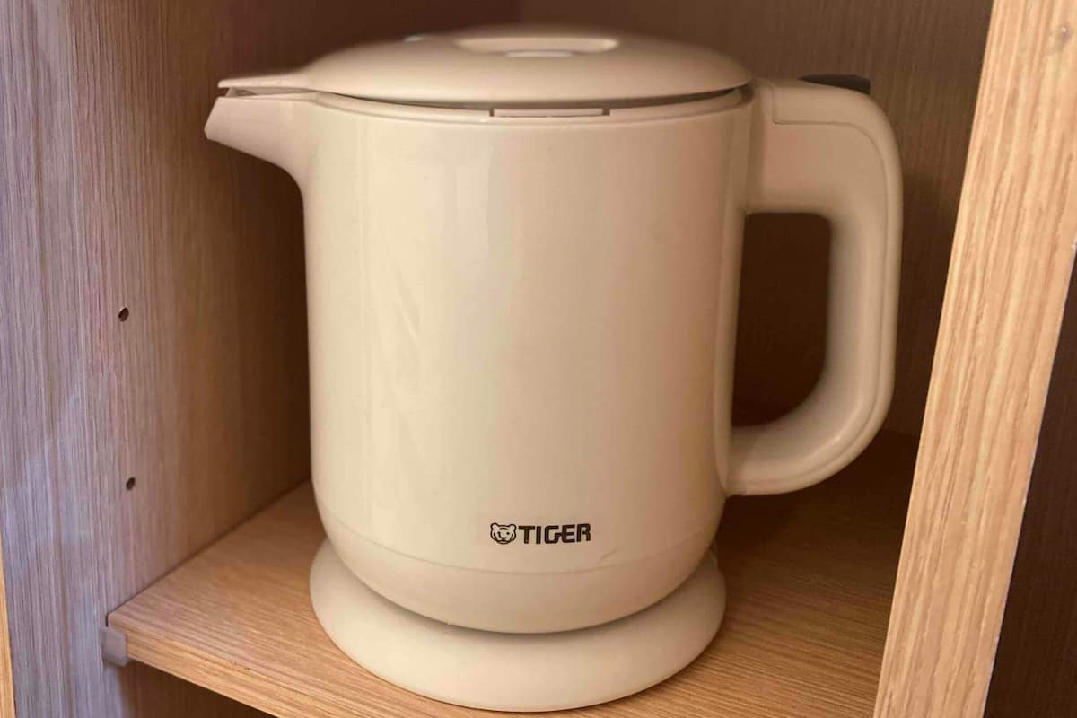 A beige electric kettle with a lid and handle, placed on a wooden shelf.