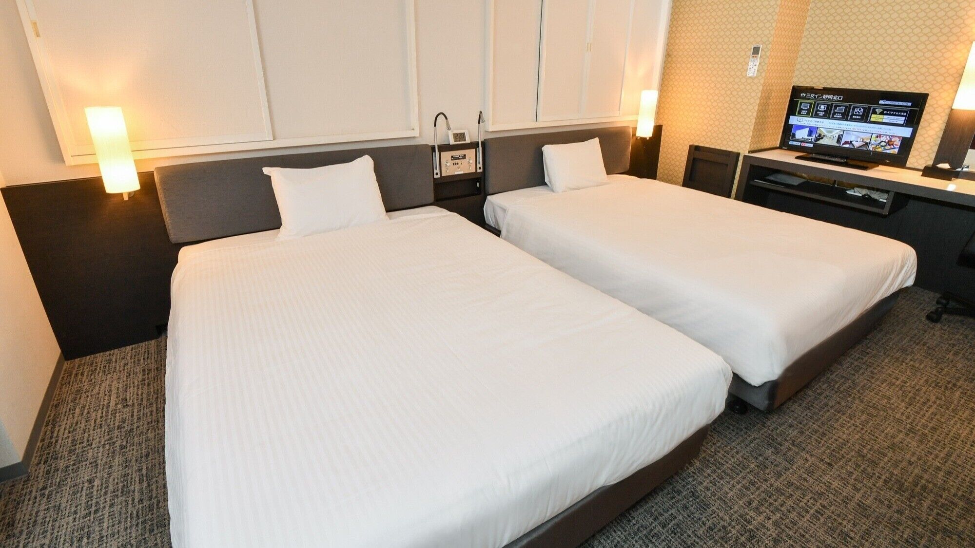 A hotel room with two beds, a desk, a television, and a wall-mounted control panel.