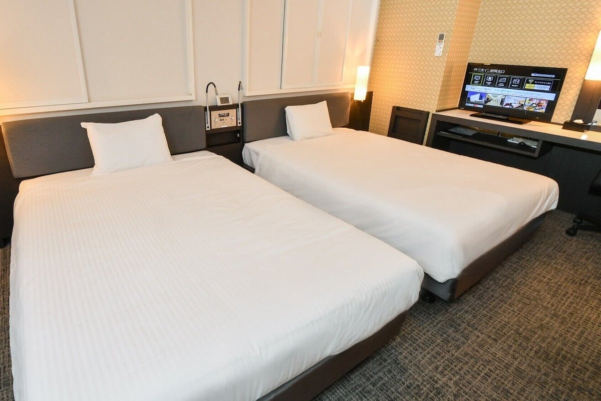 A hotel room with two beds, a desk, a television, and a wall-mounted control panel.