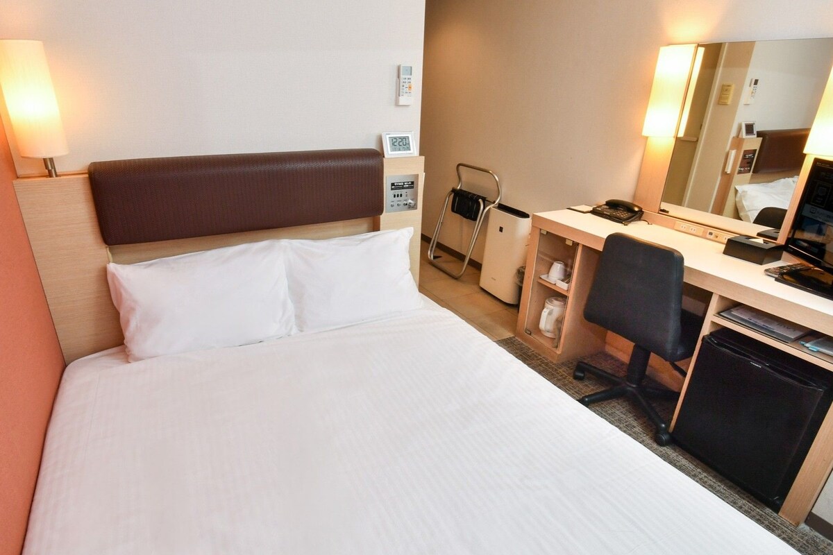 Una habitación de hotel con cama, escritorio, silla, espejo y un pequeño basurero.
