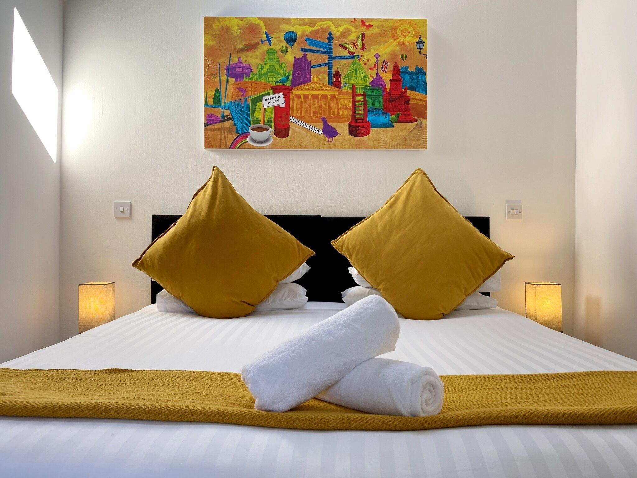 A hotel room with a bed, two mustard-colored pillows, a rolled white towel, a colorful painting on the wall, and two bedside lamps.