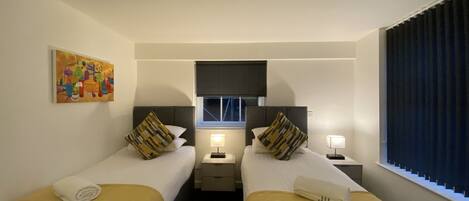 A hotel room with two beds, a painting on the wall, a window with curtains, and a ceiling light.