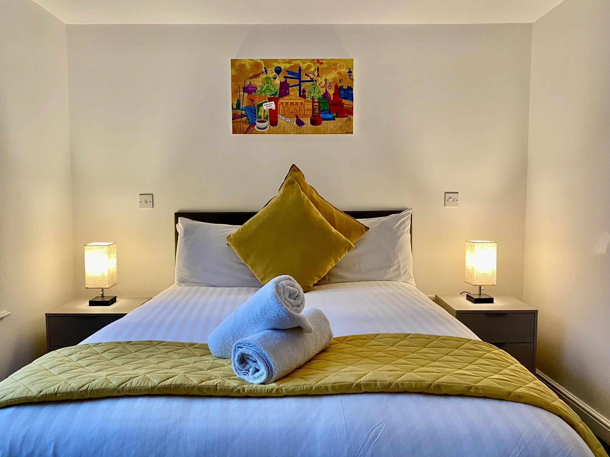 A hotel room with a bed, two bedside lamps, a painting on the wall, and a rolled towel on the bed.