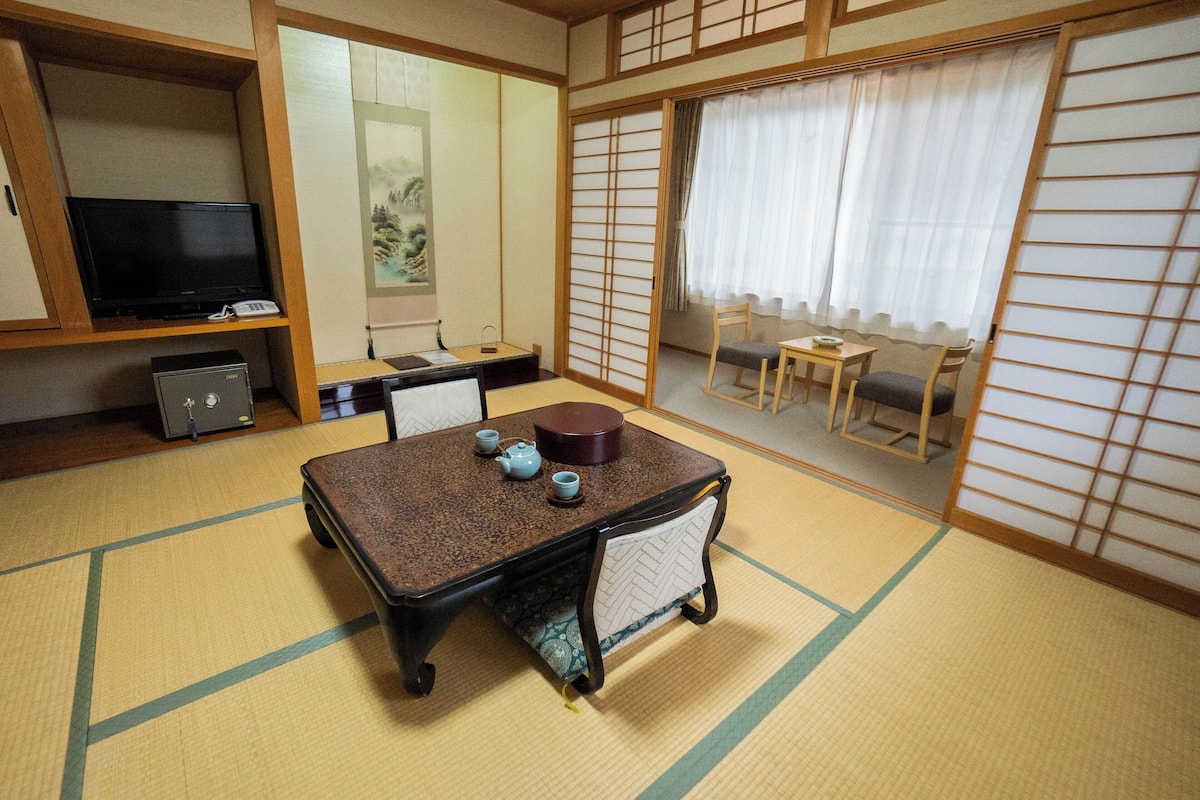 Una habitación tradicional japonesa con suelo de tatami, una mesa baja con juego de té, un televisor y puertas correderas.