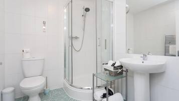 Un baño moderno con ducha de vidrio, inodoro, lavamanos y un estante pequeño con artículos de aseo.