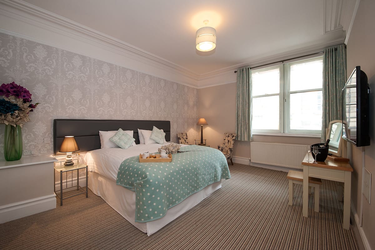 A neatly arranged bedroom with a bed, bedside lamps, a small table with a tray, and a window with curtains.