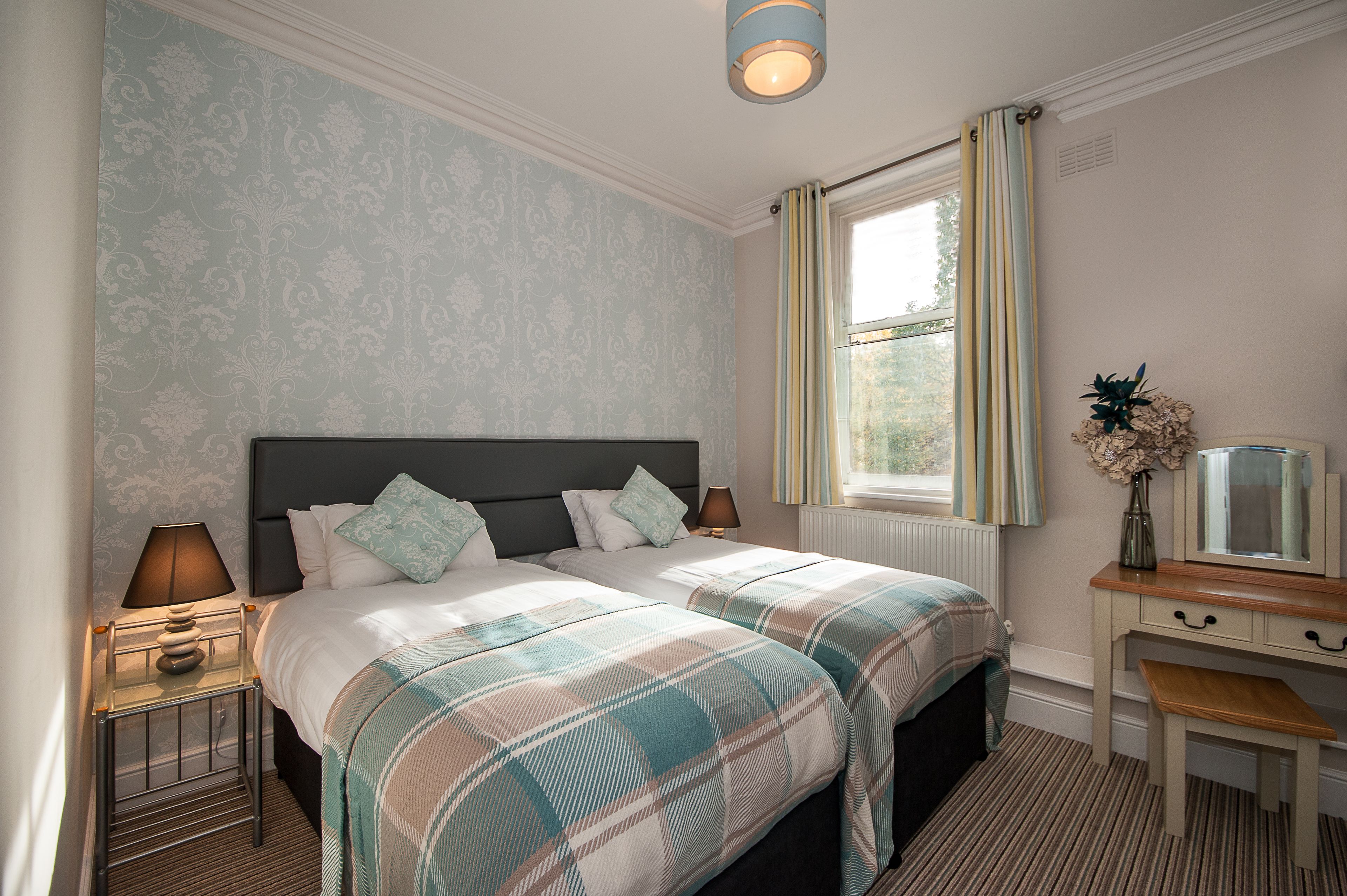 A hotel room with two beds, a desk, a lamp, and a window with curtains.
