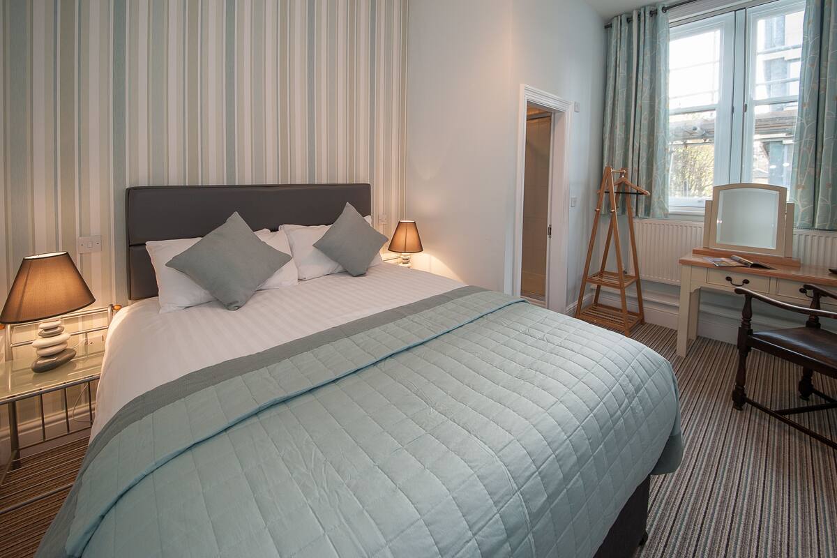 A hotel room with a bed, bedside lamps, a desk with a chair, and a window with curtains.