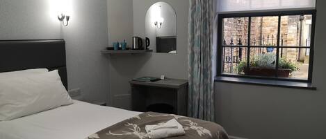A hotel room with a bed, a bedside table with a kettle, a mirror, and a window with a view of the outside.