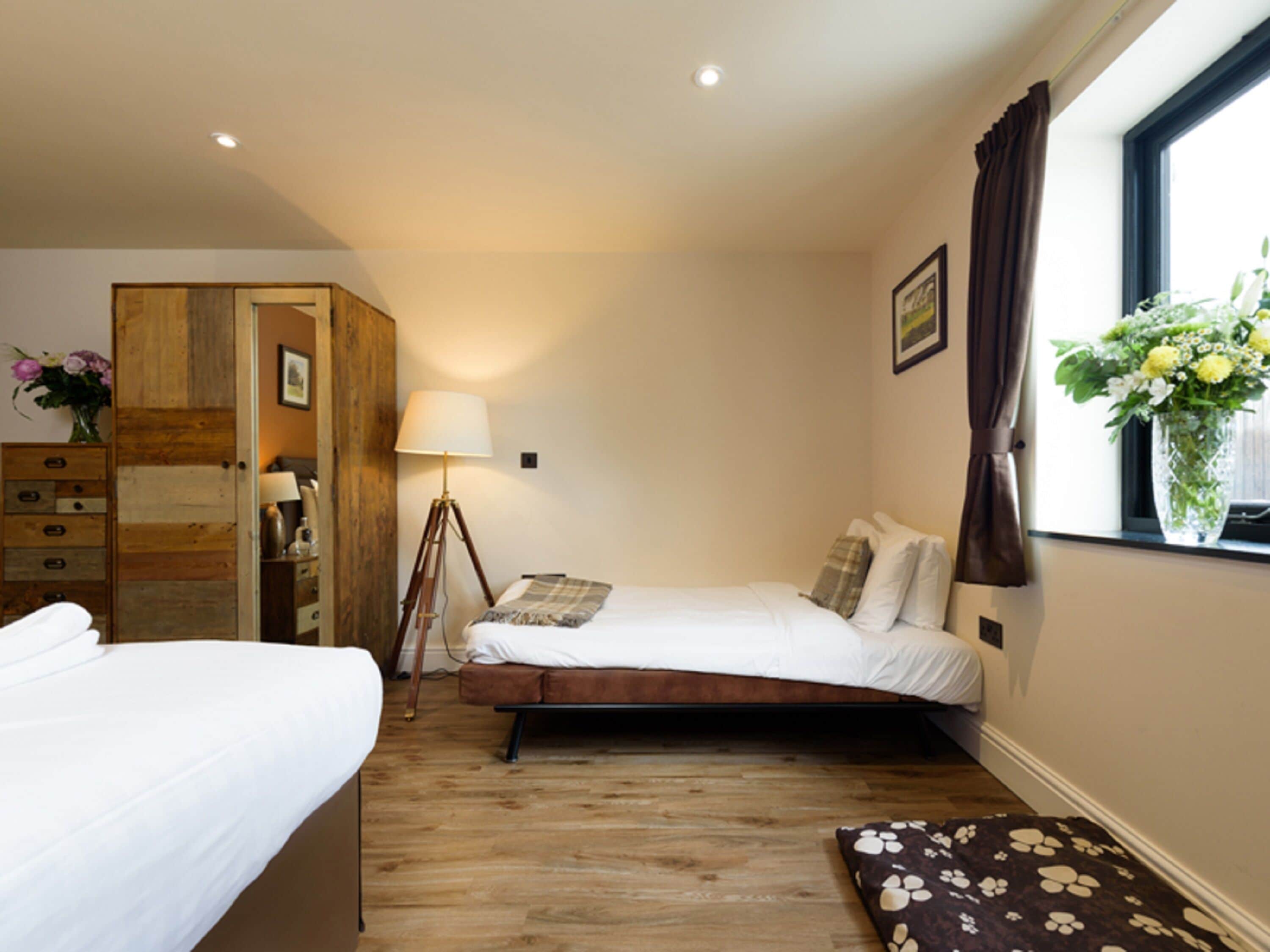 A bedroom with a wooden wardrobe, a bedside table with a lamp, a bed with a floral pillow, and a window with a view of flowers.