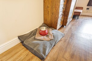 Una cama para mascotas con un cuenco sobre un piso de madera, un armario de madera y un banco marrón.